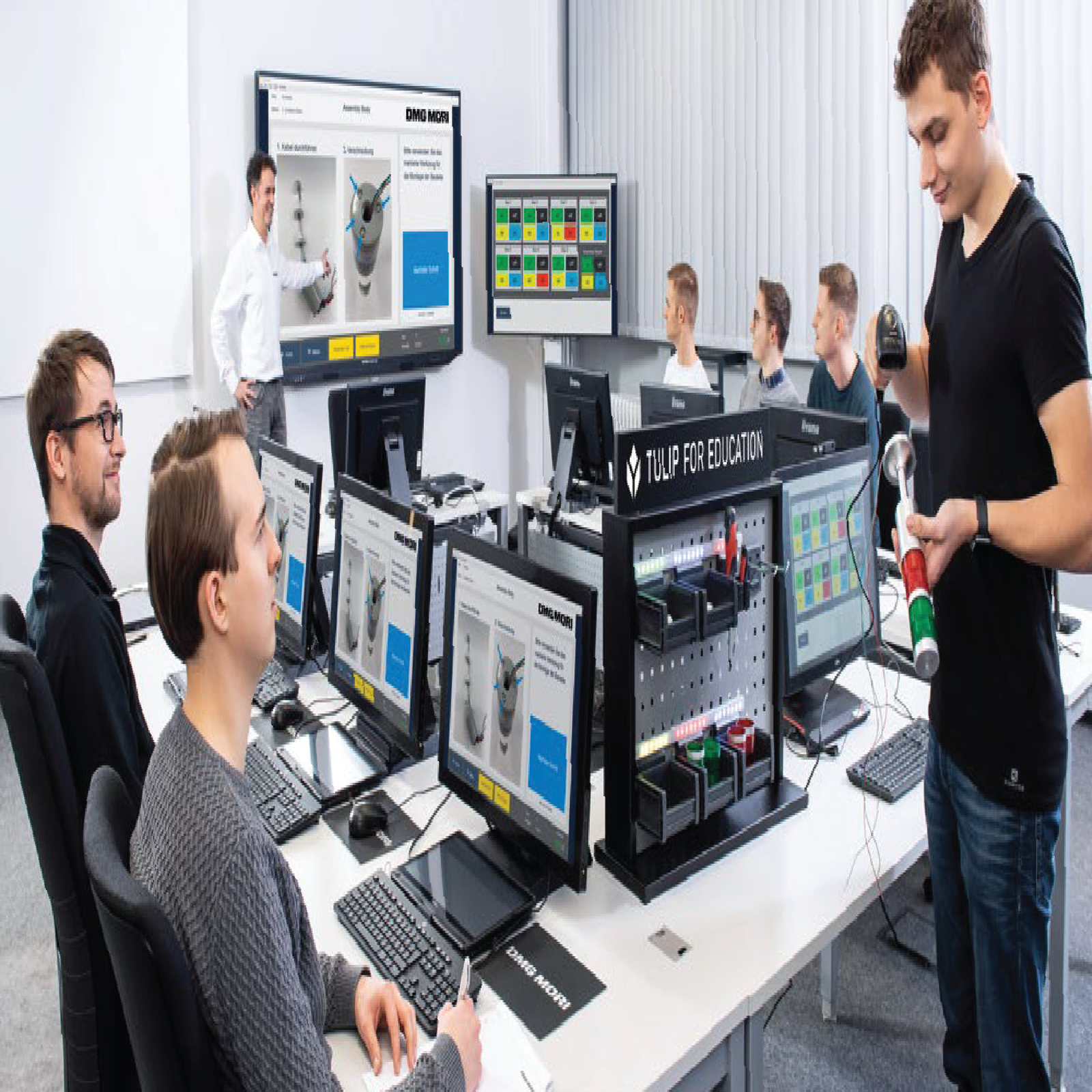 People in a modern office meeting room with glass partitions, ceiling fans, and a presenter in front of a screen